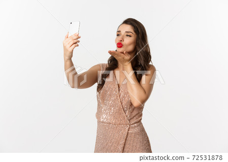 Beautiful woman with makeup and hairstyle, wearing glamour dress, taking selfie on smartphone, celebrating new year party, white background 72531878