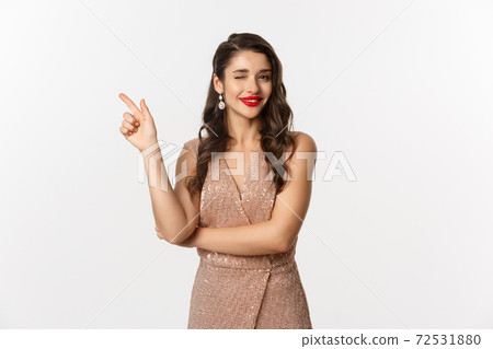 Christmas, holidays and celebration concept. Beautiful caucasian woman in party dress and red lipstick, winking and smiling, pointing finger left at promo offer, white background 72531880