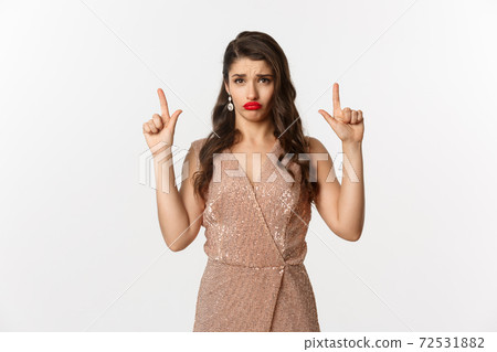 New Year, christmas and celebration concept. Gloomy and sad glamour girl in party dress pointing fingers up, sulking and crying about something, standing over white background 72531882