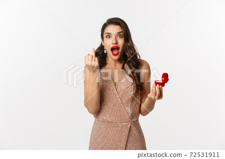 Excited young woman in glamour dress being engaged, showing finger with ring and looking happy, receive marriage proposal, standing over white background 72531911