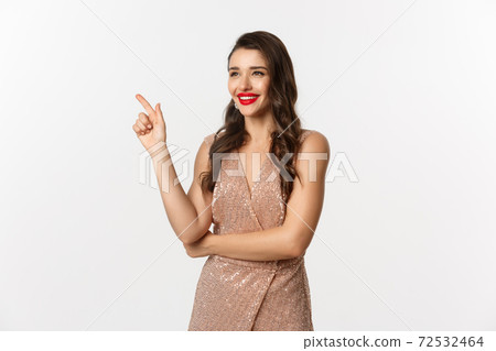 Christmas, holidays and celebration concept. Attractive woman wearing party dress and red lipstick, pointing and looking left, enjoing new year eve, white background Christmas, holidays and celebration concept. Attractive woman wearing party dress and red lipstick, pointing and looking left, enjoing new year eve, white background 72532464