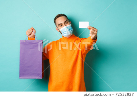 Covid-19, pandemic and lifestyle concept. Happy male customer in face mask showing credit card and purple shopping bag, standing over light blue background 72532927