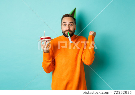 Happy birthday guy celebrating, wearing party hat, blowing wistle and holding bday cake and doing fist pump excited, standing over turquoise background Happy birthday guy celebrating, wearing party hat, blowing wistle and holding bday cake and doing fist pump excited, standing over turquoise background 72532955