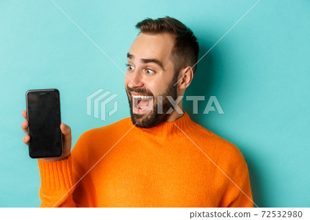 Close-up of young bearded man showing phone screen and looking amazed, wearing orange sweater, standing against studio background 72532980