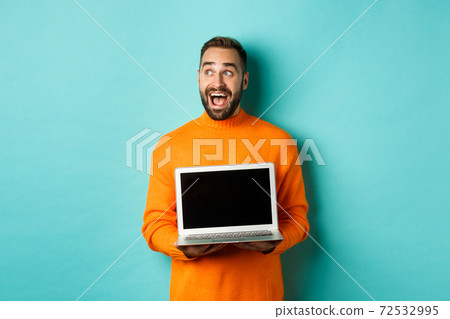 Amazed young man in orange sweater looking at upper left corner, showing laptop screen promo offer, standing over turquoise background Amazed young man in orange sweater looking at upper left corner, showing laptop screen promo offer, standing over turquoise background 72532995