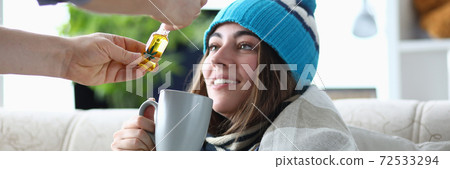 Young woman addict sits in hat and scarf on sofa at home with cup with marijuana extract portrait Young woman addict sits in hat and scarf on sofa at home with cup with marijuana extract portrait 72533294