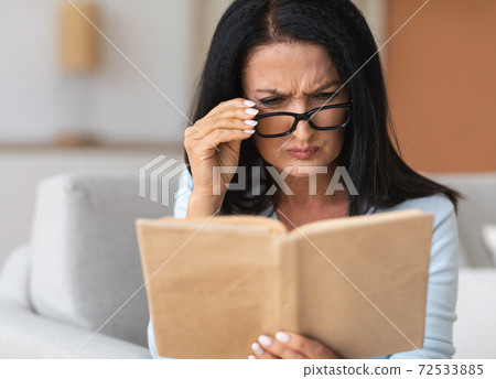 Senior confused woman in glasses trying to read book 72533885