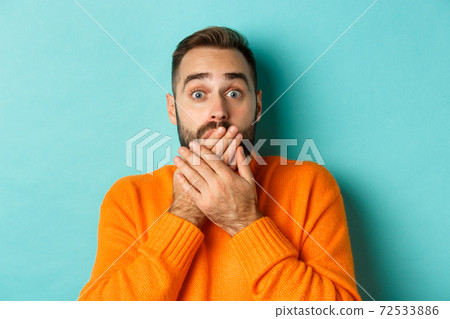 Close-up of shocked caucasian man covering his lips and mouth, staring astounded at camera, standing against turquoise background 72533886