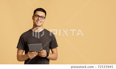Cheerful young attractive man in glasses looks at camera, holds digital tablet on online lesson 72533945