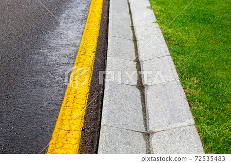 flowing rainwater in the canal is a cement ditch of a drainage system on the side of wet asphalt road with yellow markings and a green lawn on roadside environment. 72535483