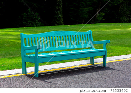 A blue wooden bench lit by sunlight stands on an asphalt road with yellow markings in a park with a green lawn and large trees in the background. A blue wooden bench lit by sunlight stands on an asphalt road with yellow markings in a park with a green lawn and large trees in the background. 72535492