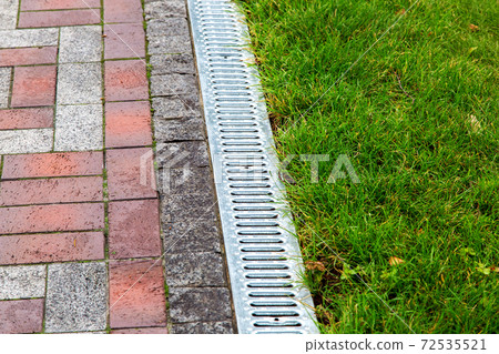 An iron gutter with grate to the drainage system on the side of the walkway with green lawn, close up. 72535521