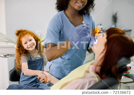 Young little red haired curly girl supporting her mom at dentist's office during checkup. Female smiling African pediatric dentist in blue uniform and gloves makes teeth filling with UV lamp Young little red haired curly girl supporting her mom at dentist's office during checkup. Female smiling African pediatric dentist in blue uniform and gloves makes teeth filling with UV lamp 72536672