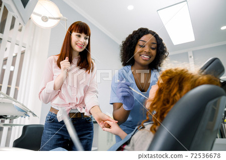 Young woman mom supporting her little daughter at dentist's office during checkup. Female smiling African pediatric dentist in blue uniform and gloves examining a little girl teeth 72536678