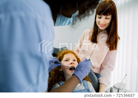 Back view of African female pediatric dental specialist making oral checkup of little curly red haired school girl, visiting dentist with her young mom. Dentist office, pediatric dentistry Back view of African female pediatric dental specialist making oral checkup of little curly red haired school girl, visiting dentist with her young mom. Dentist office, pediatric dentistry 72536687