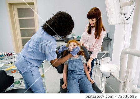 Young female African American dentist with dental tools, making dental check up of her little patient girl. Young mother reassuring her frightened cute daughter during examination 72536689