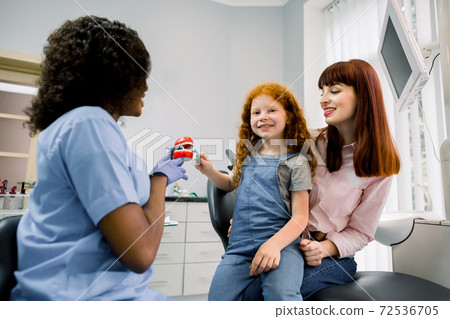 Oral care and teeth hygiene. Female African pediatric dentist explaining to young girl patient and her mother procedure of proper cleaning of teeth. Smiling child looks at camera while brushes teeth Oral care and teeth hygiene. Female African pediatric dentist explaining to young girl patient and her mother procedure of proper cleaning of teeth. Smiling child looks at camera while brushes teeth 72536705