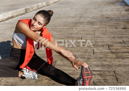 Outdoor shot of attractive fitness woman looking pleased, stretching her leg before workout, jogging on seaside promenade Outdoor shot of attractive fitness woman looking pleased, stretching her leg before workout, jogging on seaside promenade 72536998