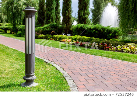 ground decorative iron lantern in the park with footpath of red stone tiles and landscaping with flower beds and bushes with trees on a sunny summer day. 72537246