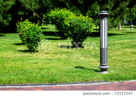 ground iron lantern in a park on a green lawn with green bushes and trees at a pedestrian footpath of red tile with a drainage system. 72537247