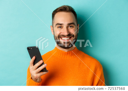 Close-up of happy handsome man writing message on mobile phone, holding smartphone and smiling, standing against turquoise background Close-up of happy handsome man writing message on mobile phone, holding smartphone and smiling, standing against turquoise background 72537568
