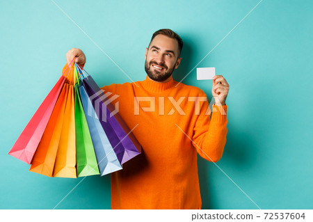 Happy aduly man showing credit card and shopping bags, standing against turquoise background Happy aduly man showing credit card and shopping bags, standing against turquoise background 72537604