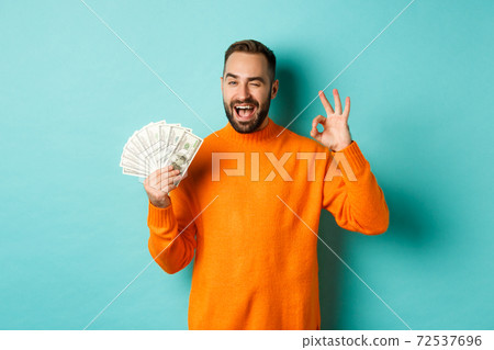 Shopping. Excited guy holding money, showing ok sign and winking, standing over light blue background Shopping. Excited guy holding money, showing ok sign and winking, standing over light blue background 72537696
