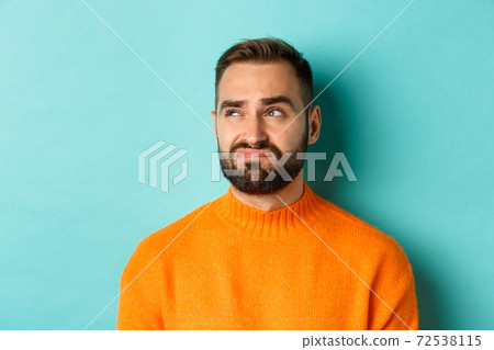 Close-up of handsome caucasian man looking left disappointed and skeptical, staring at logo, wearing orange sweater, standing against turquoise background Close-up of handsome caucasian man looking left disappointed and skeptical, staring at logo, wearing orange sweater, standing against turquoise background 72538115