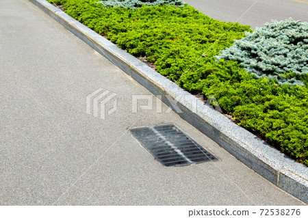 asphalt road with grate drainage system of the sewage hatch near the dividing line with plants. 72538276