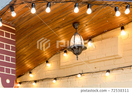 iron hanging on the ceiling retro lantern with a light bulb on the facade of a building decorated with a garland with light bulbs, close-up. 72538311