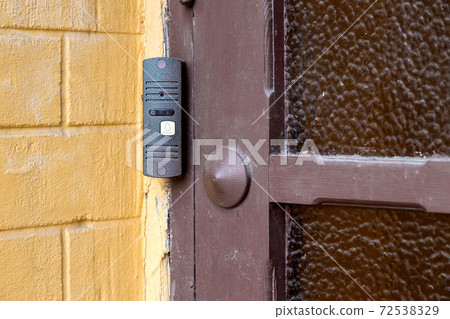 intercom with a doorbell and video link, closeup of a security device on a yellow brick wall near a wooden door with glass on outdoor nobody. 72538329