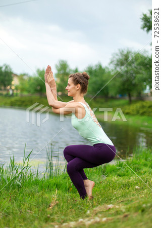 Slender Caucasian brunette girl doing yoga in summer on a green lawn by the river. Eagle Pose, Garudasana 72538621