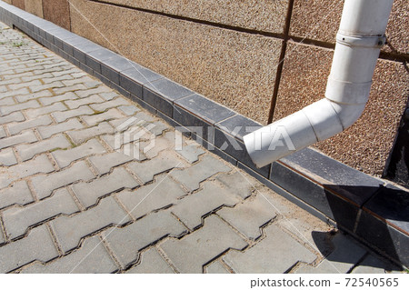 building roof drainage rain pipe with granular tar pavement wall and stone paving slabs, close-up of storm pipe on the building facade in sunny weather. 72540565