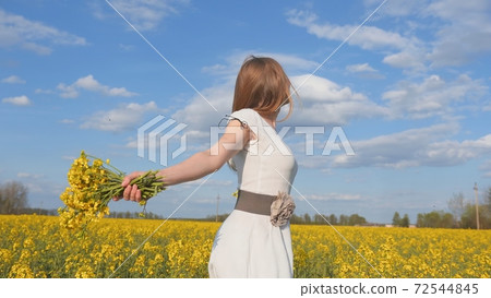 Happy girl in a white dress with a bouquet of rapeseed flowers in a yellow rapeseed field. 72544845