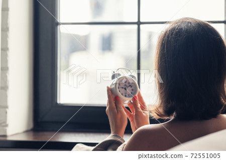Back view of lonely european young woman wrapped in a plaid with with alarm clock sitting near window at cold winter day. Back view of lonely european young woman wrapped in a plaid with with alarm clock sitting near window at cold winter day. 72551005