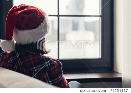 Back view of lonely european young woman wrapped in a plaid with with alarm clock sitting near window at cold winter day. 72551117