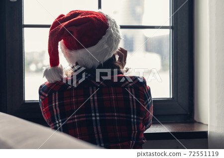 Back view of lonely european young woman wrapped in a plaid with with alarm clock sitting near window at cold winter day. 72551119