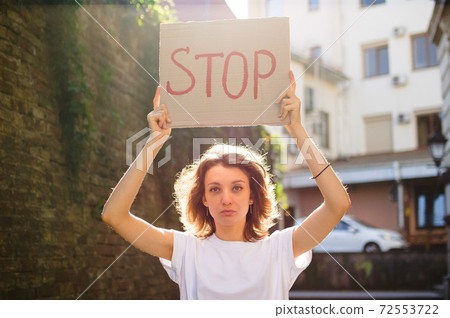 Young protesting woman in white shirt and jeans holds protest sign broadsheet placard with slogan 'Stop' for public demonstration on wall background. Young protesting woman in white shirt and jeans holds protest sign broadsheet placard with slogan 'Stop' for public demonstration on wall background. 72553722