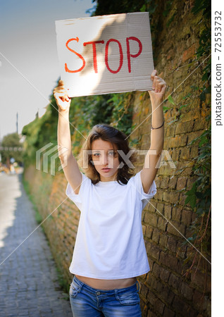 Young protesting woman in white shirt and jeans holds protest sign broadsheet placard with slogan 'Stop' for public demonstration on wall background. 72553732