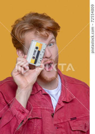 Close up of young ginger man in red jacket holding a cassette Close up of young ginger man in red jacket holding a cassette 72554809