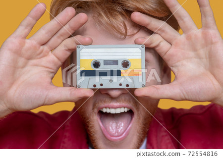 Close up of young ginger man in red jacket holding a cassette Close up of young ginger man in red jacket holding a cassette 72554816