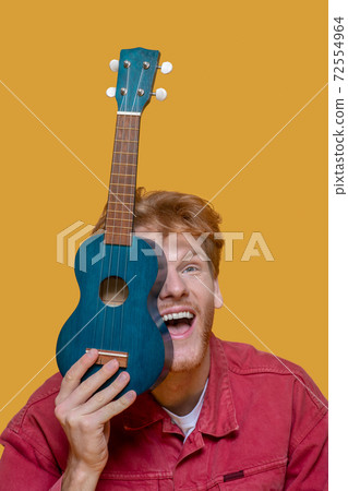 Young bearded good-looking man in red jacket with ukulele having fun Young bearded good-looking man in red jacket with ukulele having fun 72554964