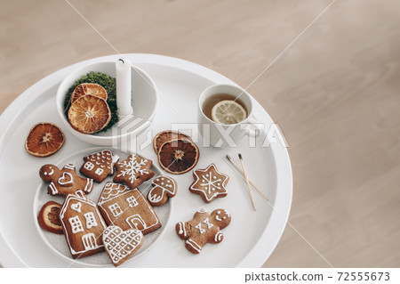 Christmas breakfast. Winter still life scene. Cup of tea, lemon and gingerbread cookies. White round coffee table. Candleholder with moss and dry orange fruit slices. Blurred wooden floor background. 72555673