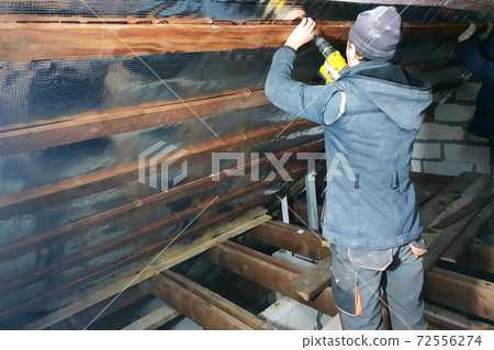 The wizard uses a cordless drill and screw the screws into boards that hold foil film and mineral wool to heal the roof of the house 72556274
