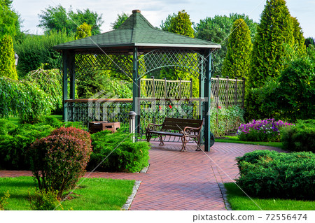 an iron gazebo with a roof and a park bench with green plants, bushes and trees on a summer day. 72556474