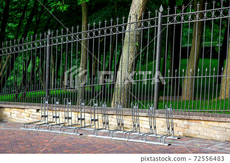 free bike parking on the sidewalk made of tiles near a black metal fence with peaks in a park with trees. free bike parking on the sidewalk made of tiles near a black metal fence with peaks in a park with trees. 72556483
