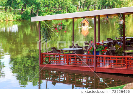floating restaurant on a red pier with a canopy over tables with chairs and flowerpots on the banks of the river with green trees on a summer day. floating restaurant on a red pier with a canopy over tables with chairs and flowerpots on the banks of the river with green trees on a summer day. 72556496