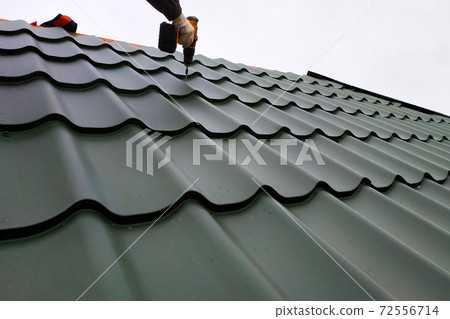 The professional worker works on installation of a roof of a roof by sheets of a metal tile and drills a screw with a drill 72556714