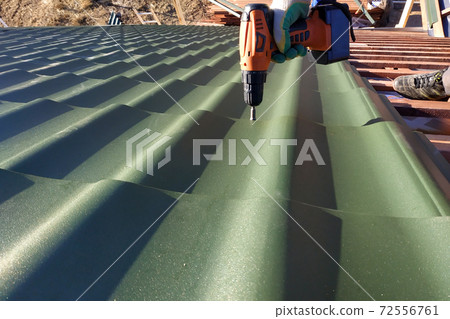 The professional worker works on installation of a roof of a roof by sheets of a metal tile and drills a screw with a drill The professional worker works on installation of a roof of a roof by sheets of a metal tile and drills a screw with a drill 72556761