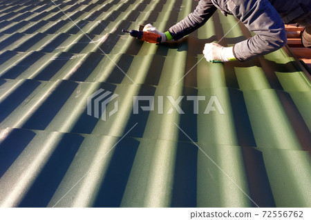 The professional worker works on installation of a roof of a roof by sheets of a metal tile and drills a screw with a drill The professional worker works on installation of a roof of a roof by sheets of a metal tile and drills a screw with a drill 72556762